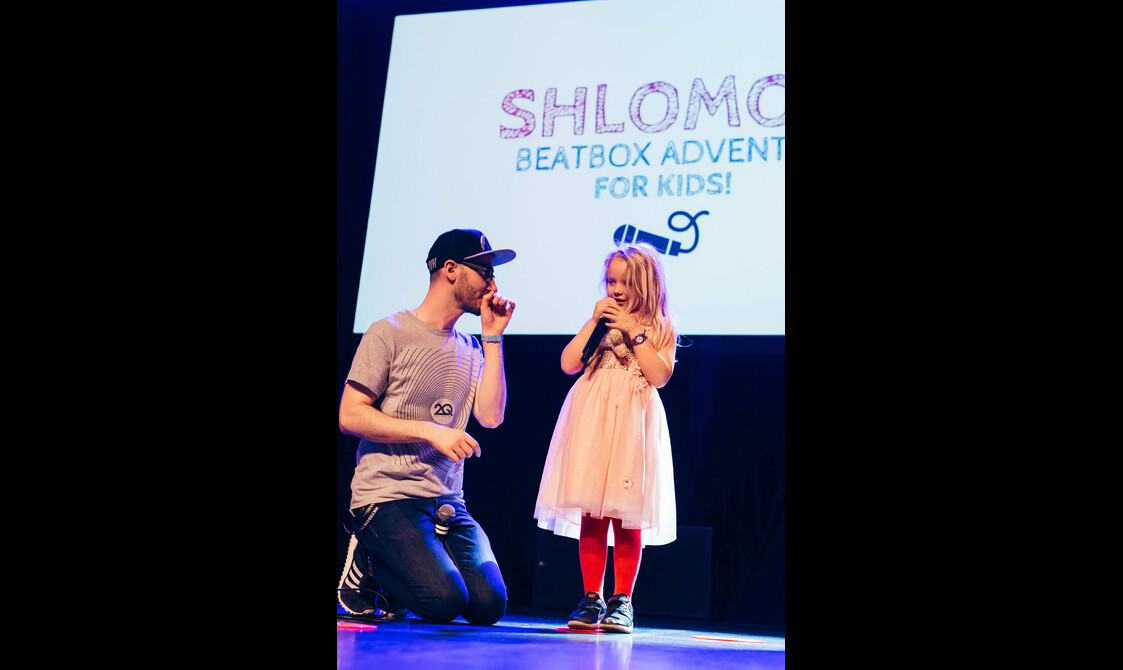 A man kneels down next to a young girl standing on stage with a microphone