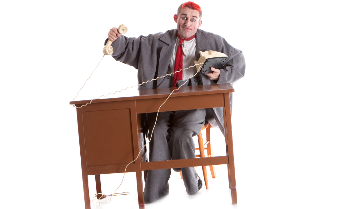 A man with red hair shrugs playfully while sitting at a wooden desk holding a knotted rotary phone.