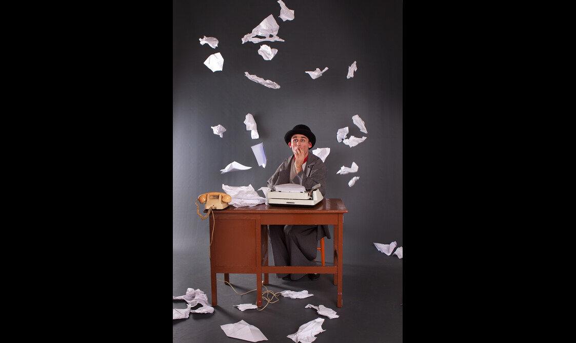 A man in a black bowler hat sits at a desk looking confused as papers fly around him.