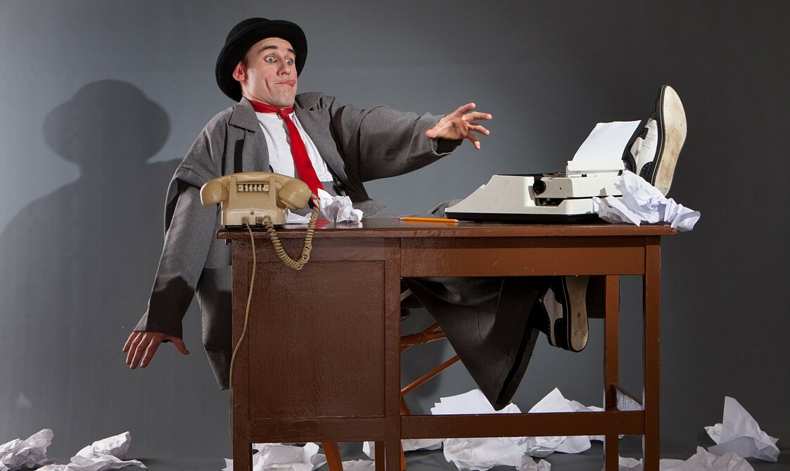 A man in a suit and black bowler hat playfully reaches forward sat at a desk towards the paper that's thrown everywhere.