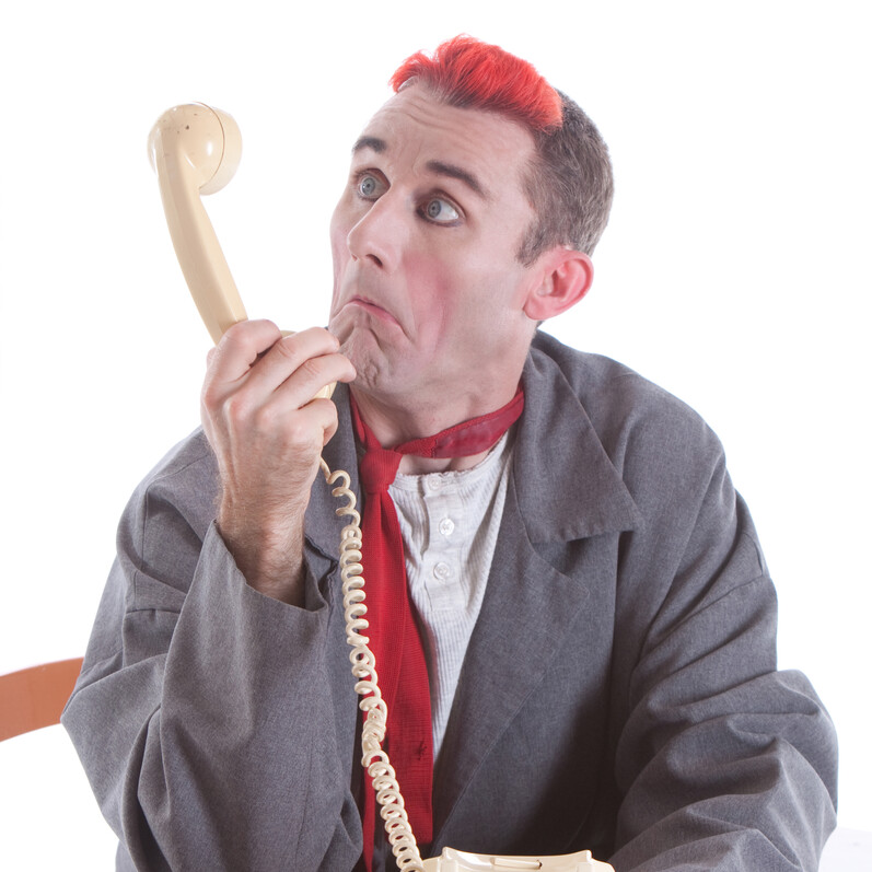 A man with red hair looks playfully confused while looking into a rotary phone, sat at a desk.