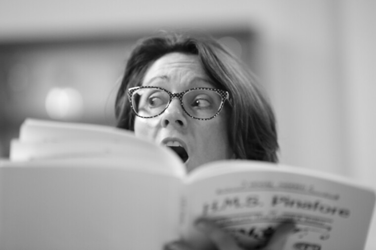 A black and white photo of a woman peering over her book looking surprised.
