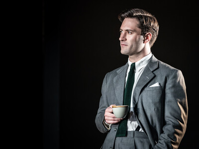 Arty Froushan stands on stage in a suit holding a cup of tea in NT lives 'Leopoldstadt'