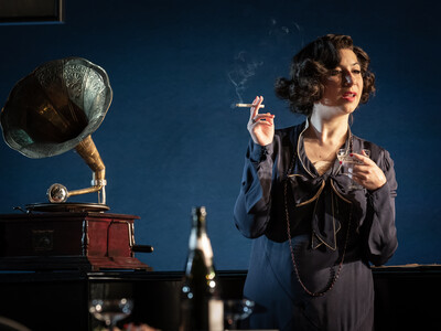 Jenna Augan smokes a cigarette and holds a glass of champagne while standing next to a gramophone in a scene from NT Lives 'Leopoldstadt'