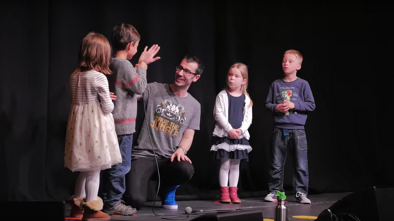 A man stands on stage with a group of children and hand fives a young boy.
