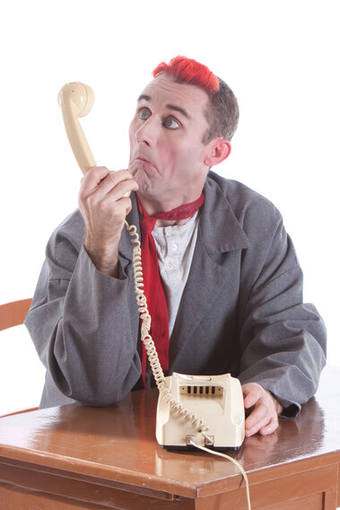A man with red hair looks playfully confused while looking into a rotary phone, sat at a desk.
