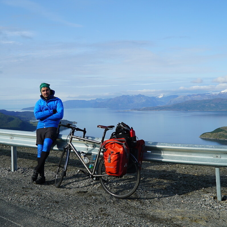 A man with folded arms, standing alongside his bike with a breathtaking view over a lake