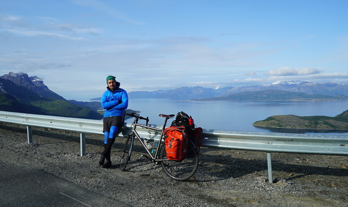 A man with folded arms, standing alongside his bike with a breathtaking view over a lake