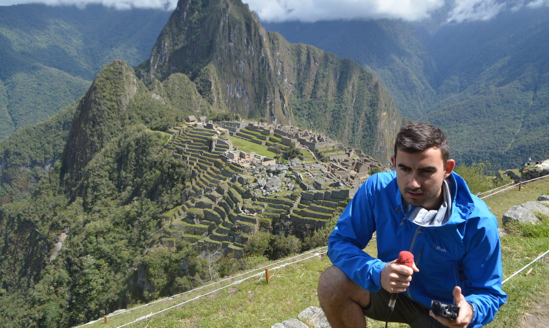 A man, crouching with a microphone in his hand with a view of mountains in the background