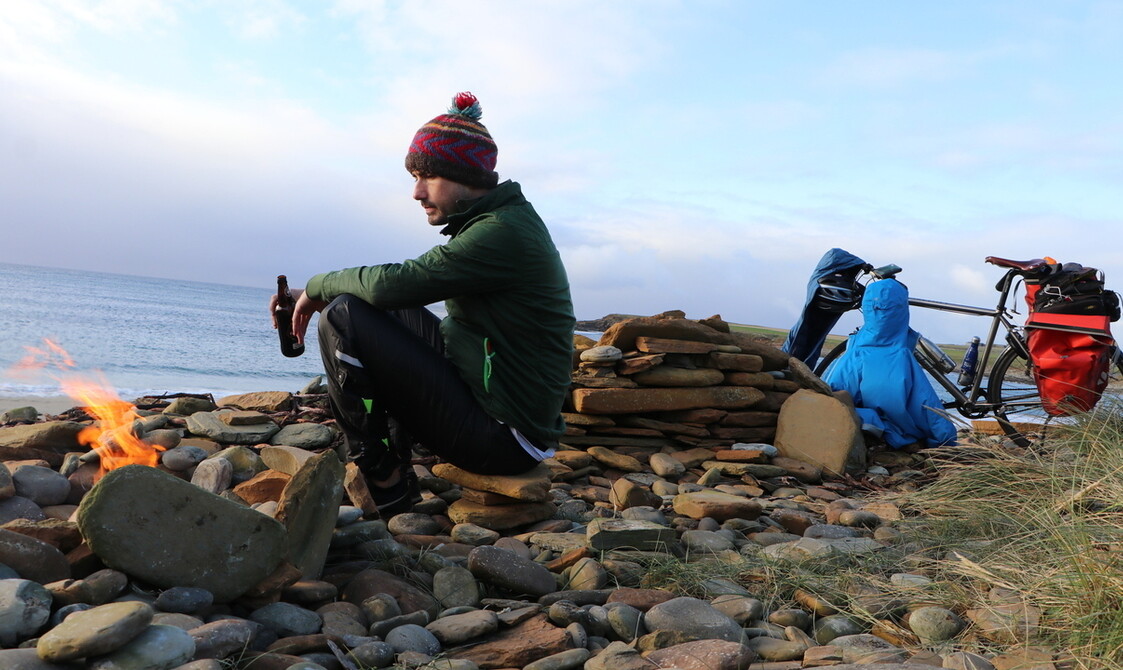 A man wearing a bobble hat, sitting with a bottle in his hand, beside an open fire on the edge of a lake
