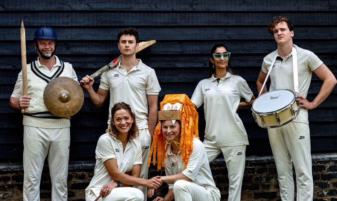 6 actors wearing cricket whites stand and crouch in front of a building. Some hold or wear props.