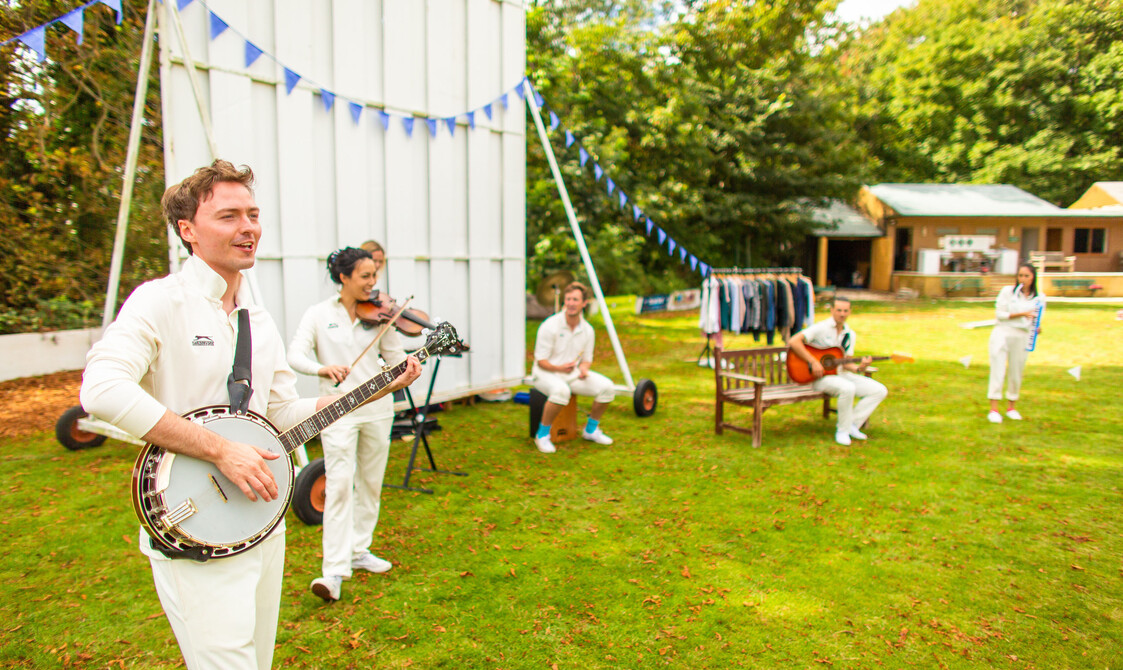 6 actors wearing cricket whites playing various instruments on a cricket green