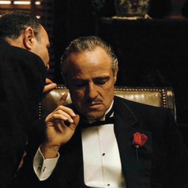 A man whispers into the ear of a man wearing a black tuxedo that has a red rose in the button hole