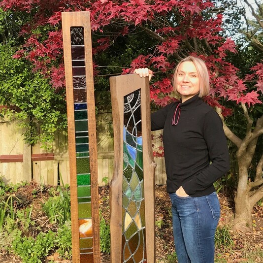 A blonde woman stands in a garden with her arm resting on one of two glass mosaic sculptures.
