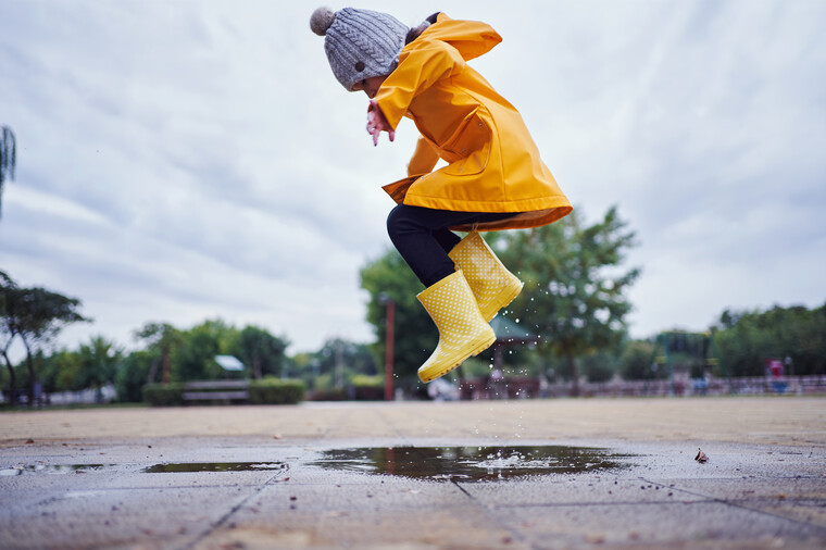 Young child in a yellow rain jacket and wellies jumping in a puddle.