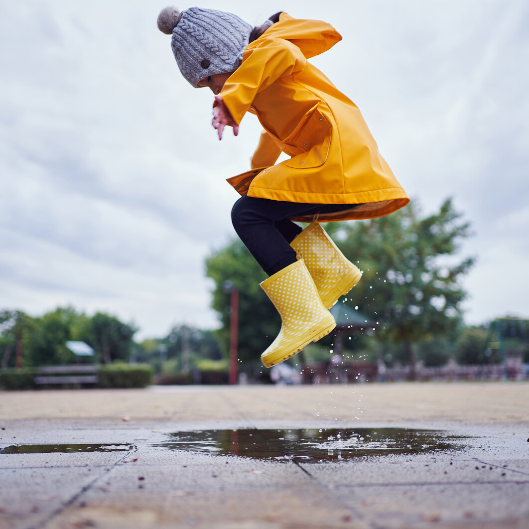 Young child in a yellow rain jacket and wellies jumping in a puddle.