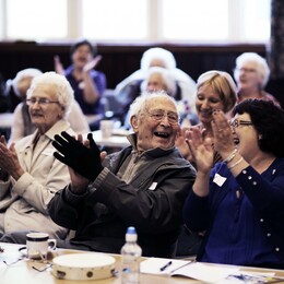 An elderly audience claps in a hall.