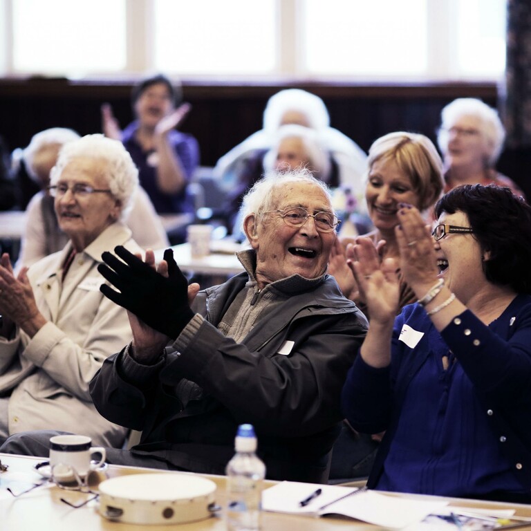 An elderly audience claps in a hall.
