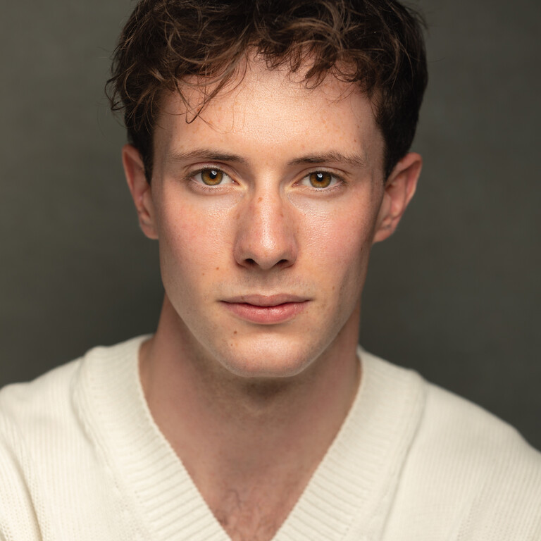 Headshot of a young man with dark hair, wearing a white v-neck jumper