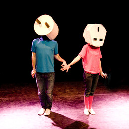 Two young people stand holding hands on stage with large plug/charger sculptures over their faces
